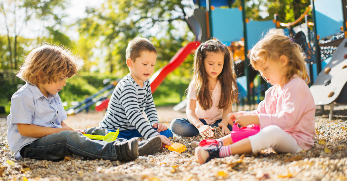 Four preschool children sitting on the ground and exploring rocks and natural materials with small shovels and cups in the playground
