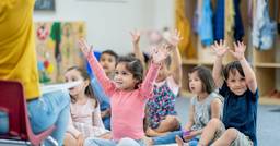 Group of preschoolers sitting on the carpet during circle time looking at their teacher with their arms raised in the air 