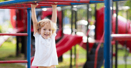 A preschool boy hanging on red monkey bars and smiling at the playground