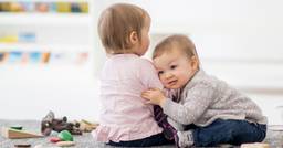 Two infants, one facing the camera and one with their back to the camera, hugging in their infant classroom