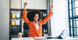 Childcare director sitting at her office desk, smiling at her computer with her arms up in the air. 